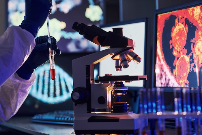 scientist-in-white-protective-uniform-works-with-c-2026-03-24-03-20-11-utc Monitors with information on the table. Scientist in white protective uniform works with coronavirus and blood tubes in laboratory.